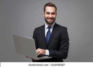 Smiling Young Business Man In Classic Black Suit Shirt Tie Posing Isolated On Grey Background. Achievement Career Wealth Business Concept. Mock Up Copy Space. Holding, Working On Laptop Pc Computer