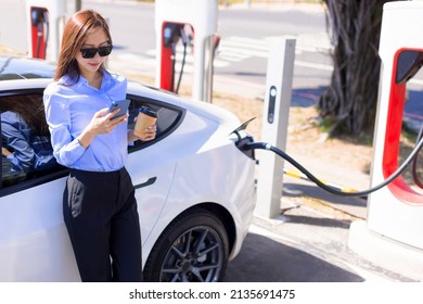 Smiling young business woman standing on city parking near electric car, charging automobile battery from small city station, drinking coffee and using smartphone - Powered by Shutterstock