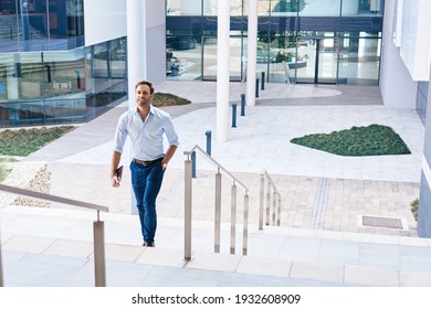 Smiling young businessman with a digital tablet walking up some stairs outside of a modern office building - Shutterstock ID 1932608909