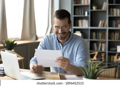Smiling young businessman in eyeglasses looking at paper correspondence, reading pleasant good news. Excited happy male entrepreneur getting bank loan approvement notification at home office. - Shutterstock ID 1896452515