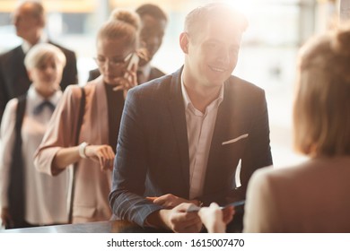 Smiling young businessman giving his ticket to the receptionist and registering on business conference with other business people in the background - Shutterstock ID 1615007470