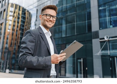 A smiling young businessman in a gray suit uses a tablet outdoors, standing in front of modern office buildings.

 - Powered by Shutterstock