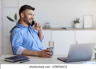 Smiling young businessman having coffee break at workplace and talking on cellphone while working on laptop in modern white office, copy space - Powered by Shutterstock