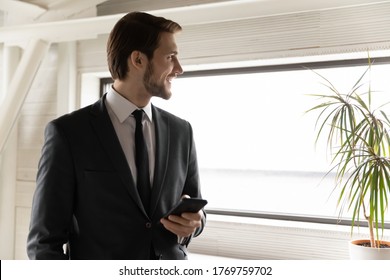 Smiling young businessman hold cellphone look in window distance thinking or visualizing, happy Caucasian male employee use smartphone, ponder plan future career success, business vision concept