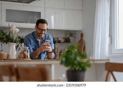 Smiling young businessman sending messages over smart phone while standing at kitchen counter - Powered by Shutterstock