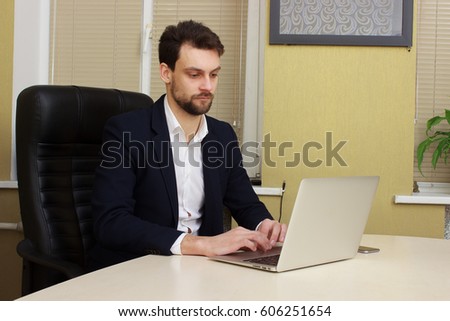 Smiling young businessman sitting behind his desk with laptop in office