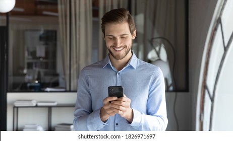 Smiling Young Businessman Standing In Modern Office, Using Smartphone Applications, Setting Meeting Reminder In Mobile Organizer Or Communicating Online With Clients, People And Technology Concept.