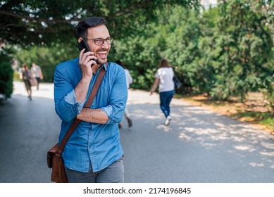 Smiling Young Businessman Talking On Smart Phone. Professional Commuter Is Carrying Laptop Bag. He Is In Formals While Walking On Road Against Trees At The City.