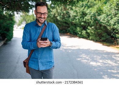 Smiling Young Businessman Text Messaging On Smart Phone. Male Professional Is Carrying Laptop Bag. He Is In Formals While Walking Against Trees.