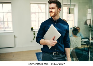 Smiling Young Businessman Walking With His Laptop In A Modern Office With Colleagues At Work In The Background