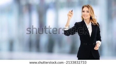 Smiling young businesswoman in black confident suit, writing or drawing on screen or transparent glass by marker, indoors. Blurred office background. Advertising sales deal offer. Save big concept.