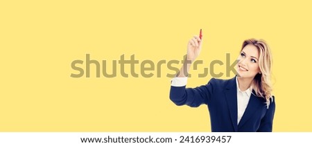 Smiling young businesswoman in blue confident suit, writing or drawing some ad text on screen transparent glass, by red marker, isolated against yellow background. Wide banner image.