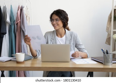 Smiling young businesswoman designer in eyeglasses holding market research paper document, analyzing charts data sales of personal fashion clothing brand, working on computer alone in modern office. - Powered by Shutterstock