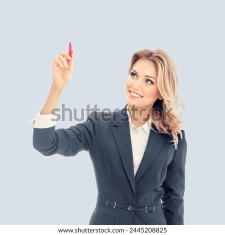 Smiling young businesswoman in grey confident suit, writing or drawing something on screen or transparent glass, by marker, isolated against gray background. Square composition image.