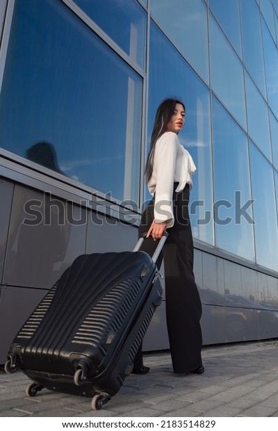 Smiling young businesswoman passes airport\
terminal with suitcase in hand. Brunette with long hair in formal\
blouse and trousers travels at\
sunset
