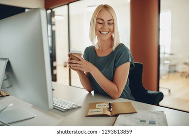 Smiling young businesswoman sitting at her desk in an office reading paperwork and drinking a cup of takeaway coffee