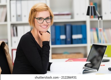 Smiling Young Businesswoman Wearing Glasses Sitting At Her Desk In The Office Doing Paperwork And Turning To Look At The Camera With A Friendly Grin