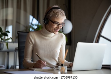 Smiling Young Businesswoman Wearing Headphones Sit At Desk Make Notes Watching Webinar On Computer, Caucasian Female Employee In Headset Write Having Video Call Conference With Colleague On Laptop