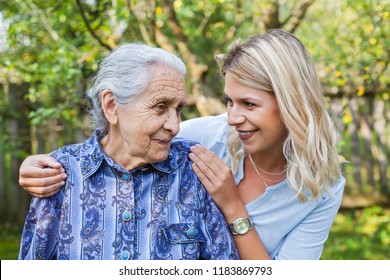 Smiling Young Caretaker Walking With Senior Lady In The Garden