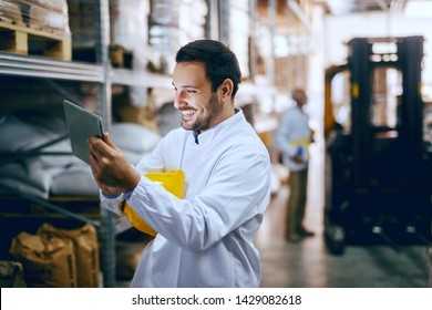 Smiling Young Caucasian Blue Collar Worker In White Uniform And With Helmet Under Armpit Using Tablet For Controling Delivery. In Background Worker Standing Next To Forklift. Warehouse Interior.