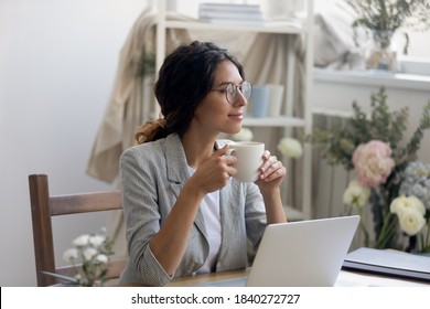 Smiling young Caucasian businesswoman in glasses drink coffee enjoy morning in home office. Happy female fashion designer stylist look in distance dreaming thinking, work in cozy creative workplace.