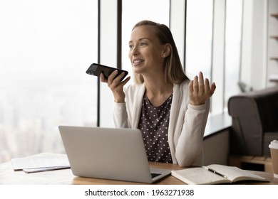 Smiling Young Caucasian Businesswoman Work On Computer In Office Talk On Loudspeaker On Smartphone. Happy Female Employee Record Audio Voice Message On Cellphone, Activate Digital Assistant.