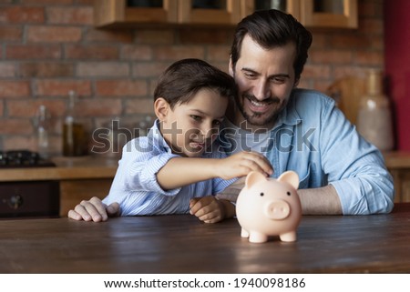 Smiling young Caucasian dad and little 7s son manage family budget put coin into piggybank save money for future needs. Happy smart father and small boy child make financial investment. Bank concept.