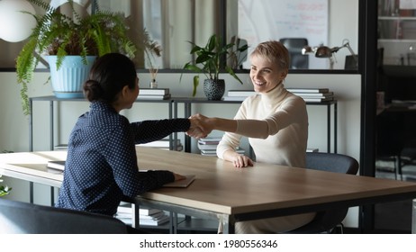 Smiling young Caucasian employee shake hand of ethnic female colleague or business partner get acquainted greet at meeting. Happy multiracial businesswomen handshake close deal make agreement. - Shutterstock ID 1980556346