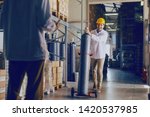 Smiling young Caucasian employee transporting bottle with gas while manager holding tablet and talking to worker. Warehouse interior.