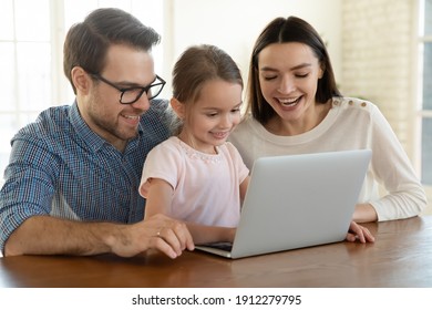 Smiling Young Caucasian Family With Small 7s Daughter Look At Laptop Screen Watching Funny Video Online. Happy Mom And Dad Have Fun Using Computer With Little Girl Child At Home. Technology Concept.