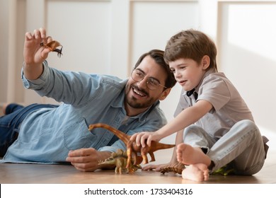 Smiling young Caucasian father and little preschooler son sit on warm floor at home play with rubber dinosaur figures together, playful dad and small boy child have fun engaged in game at home
