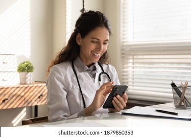 Smiling young Caucasian female doctor in white medical uniform sit at desk in hospital look at cellphone screen consult client online. Happy woman GP or physician use smartphone in modern clinic. - Powered by Shutterstock