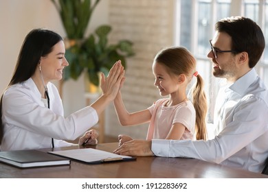 Smiling Young Caucasian Female Pediatrician Or Nurse Give High Five Make Deal With Small Girl Child Patient. Happy Woman Doctor Cheer Positive Little Kid At Hospital Visit With Dad. Medicine Concept.