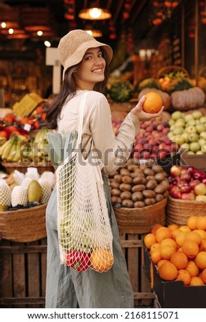 Smiling young caucasian girl is in market choosing oranges when buying fruits. Brunette wears hat, blouse, pants and string bag. Concept of natural organic products