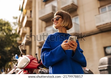 Smiling young caucasian girl with smartphone in her hands, looks away relaxing outdoors. Brown-haired woman wears sunglasses and blue sweater. Happy weekend concept, use Stock photo © 