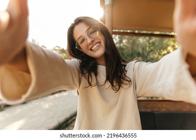 Smiling young caucasian girl takes selfie stretching hands to camera in sunny weather outdoors. Brunette wear glasses, everyday sweatshirt. Emotion concept - Powered by Shutterstock