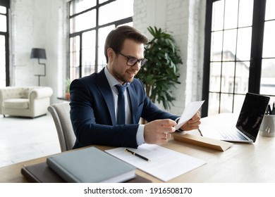 Smiling Young Caucasian Male Director Or CEO Sit At Table In Office Read Good News In Post Paper Correspondence. Happy Businessman Receive Pleasant Message In Paperwork Or Document Letter.