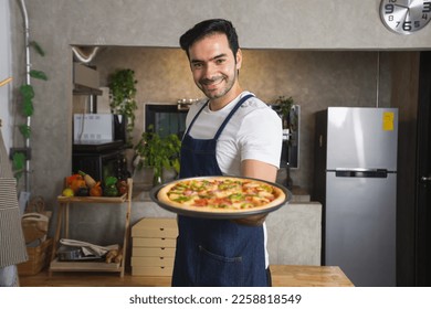 smiling young Caucasian man in apron and holding baking tray with fresh homemade pizza. Confident male chef holding cooked homemade pizza in kitchen. Italian delicious appetizer - Powered by Shutterstock