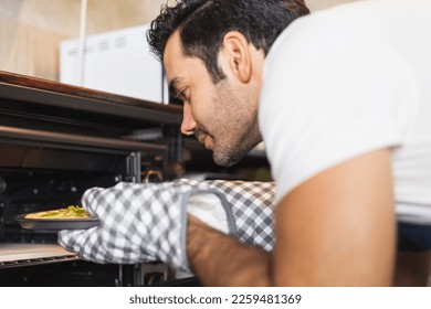 Smiling young Caucasian man in apron wearing oven mitt putting homemade pizza tray to the oven. Satisfied chef baking food at home. Italian delicious appetizer