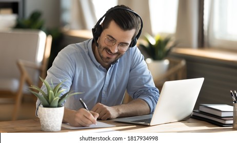 Smiling Young Caucasian Man In Headphones Glasses Sit At Desk Work On Laptop Making Notes. Happy Millennial Male In Earphones Watch Webinar Or Training Course Or Computer, Study Online From Home.