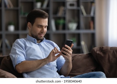 Smiling Young Caucasian Man Relax Sit On Sofa At Home Use Cellphone Gadget Texting Messaging. Happy Millennial Male Browse Wireless Internet On Smartphone Device Online. Communication Concept.