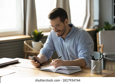 Smiling Young Caucasian Man Sit At Desk At Home Office Workplace Look At Cellphone Screen Watch Webinar Making Notes. Happy Millennial Male Use Smartphone Summarize Plan Writing In Journal.