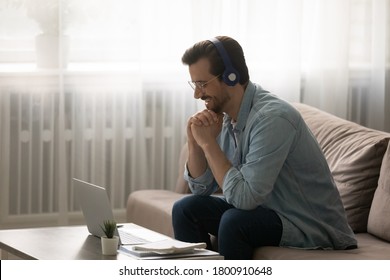 Smiling young Caucasian man in wireless headphones sit on couch at home watch webinar on laptop, happy millennial male in earphones glasses take online course training on computer, education concept - Shutterstock ID 1800910648
