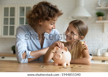 Smiling young Caucasian mom and little daughter put coins money into piggybank mange family budget together. Happy mother and small girl child saving for future feel economical provident.