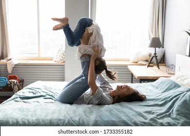 Smiling young Caucasian mom lying on comfortable bed have fun engaged in funny game activity with small daughter, overjoyed mother and little girl child playing together in cozy bedroom at home