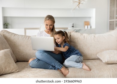 Smiling Young Caucasian Mom And Small Daughter Sit On Sofa At Home Use Modern Laptop Together. Happy Mother And Little Girl Child Relax On Couch Study Online Talk On Video Call On Computer.