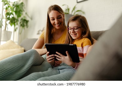 Smiling Young Caucasian Mother And Small Preschooler Daughter Sit Relax On Couch In Living Room Using Tablet. Happy Mom Or Nanny And Little Girl Child Rest On Sofa Watch Video On Tablet.