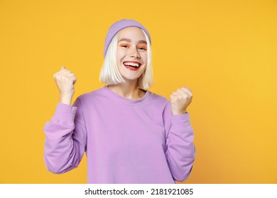 Smiling young caucasian woman 20s bob haircut bright makeup in basic purple shirt violet beanie hat looking camera doing winner gesture clenching fistd isolated on yellow background studio portrait