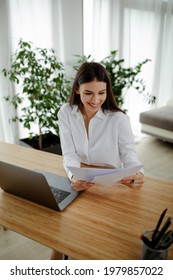 Smiling Young Caucasian Woman Holding Paper, Doing Paperwork, Reading Good News, Checking Post Mail Sitting At Home Table.