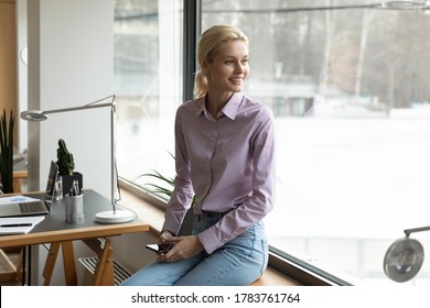 Smiling young Caucasian woman sit on windowsill look in window distance thinking or visualizing bright future career, happy female use smartphone lost in thoughts, dreaming or planning in office - Shutterstock ID 1783761764